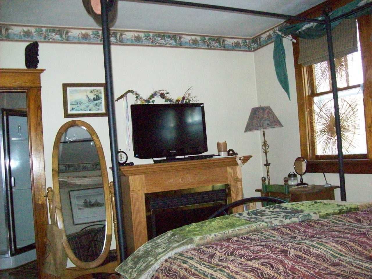 Ein Schlafzimmer mit Fernseher auf einem Holztisch, einem Spiegel, einem Bett mit gemustertem Überwurf und einem Fenster mit Jalousien.