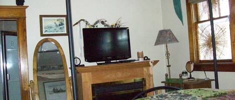 Ein Schlafzimmer mit Fernseher auf einem Holztisch, einem Spiegel, einem Bett mit gemustertem Überwurf und einem Fenster mit Jalousien.
