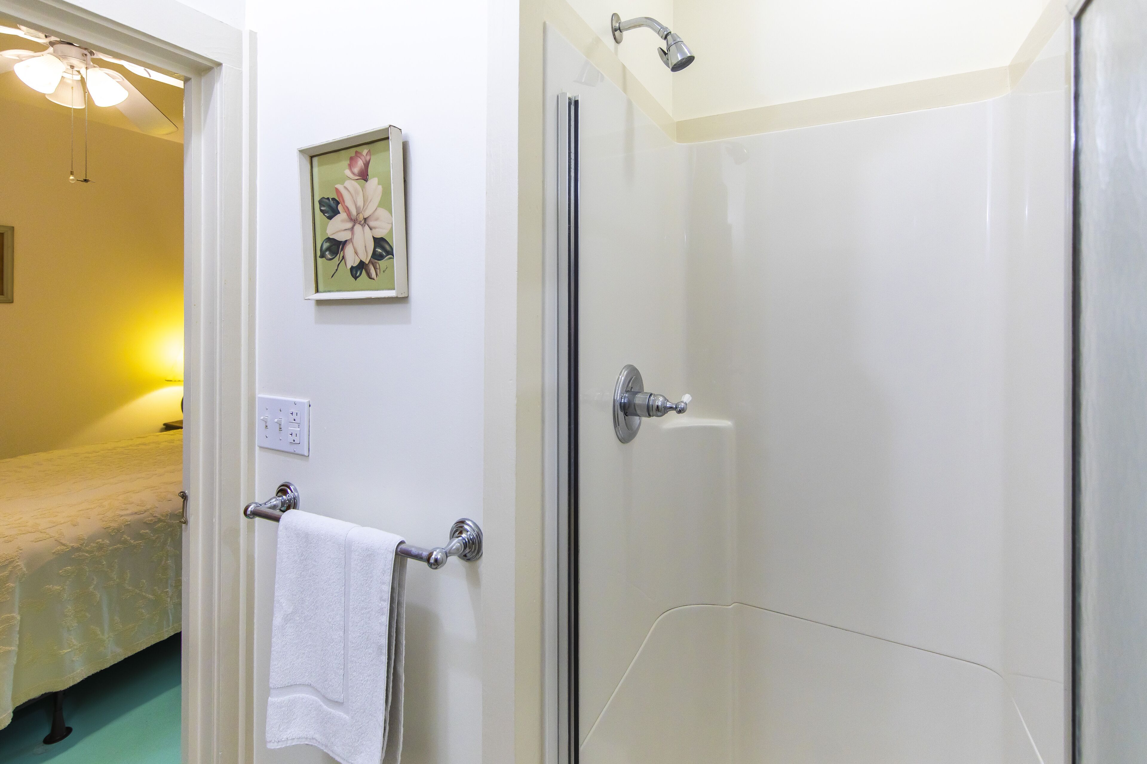 A bathroom with a glass shower door, a towel rack with a white towel, and a bed with a floral-patterned bedspread.