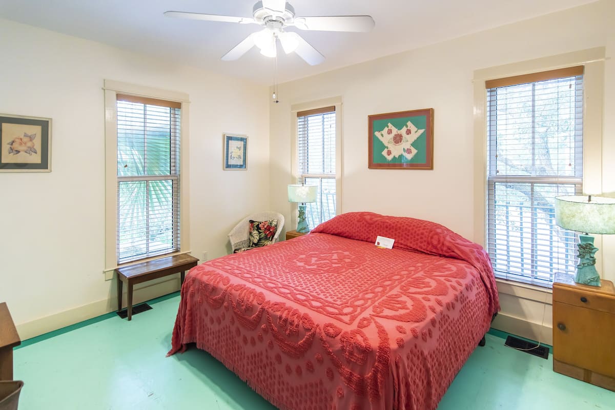 A bedroom with a large bed, a ceiling fan, and two windows with blinds.