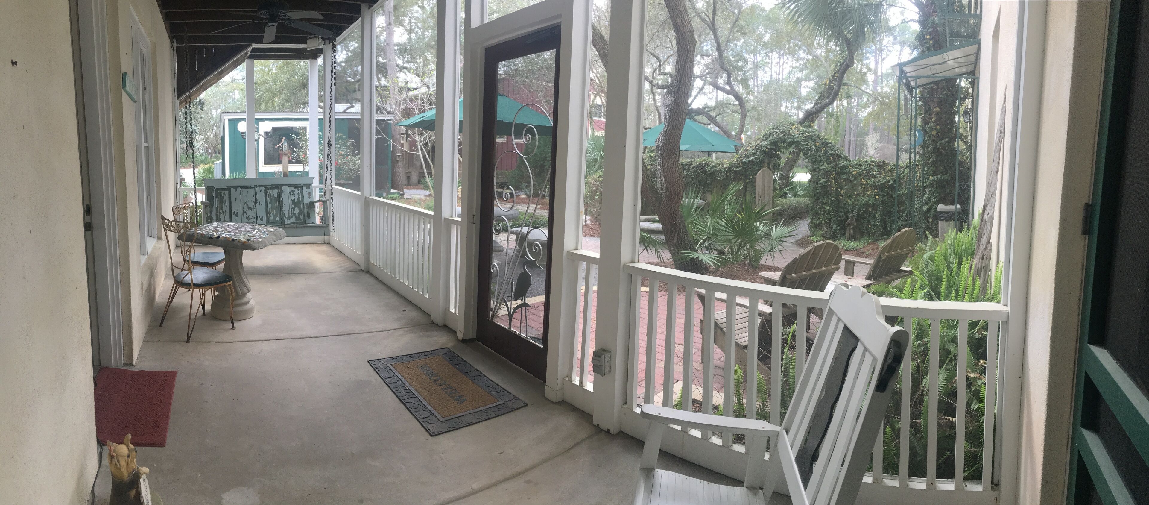 Un porche cubierto con una banca, una mesa, un felpudo y vistas a un jardín con árboles y un gazebo.