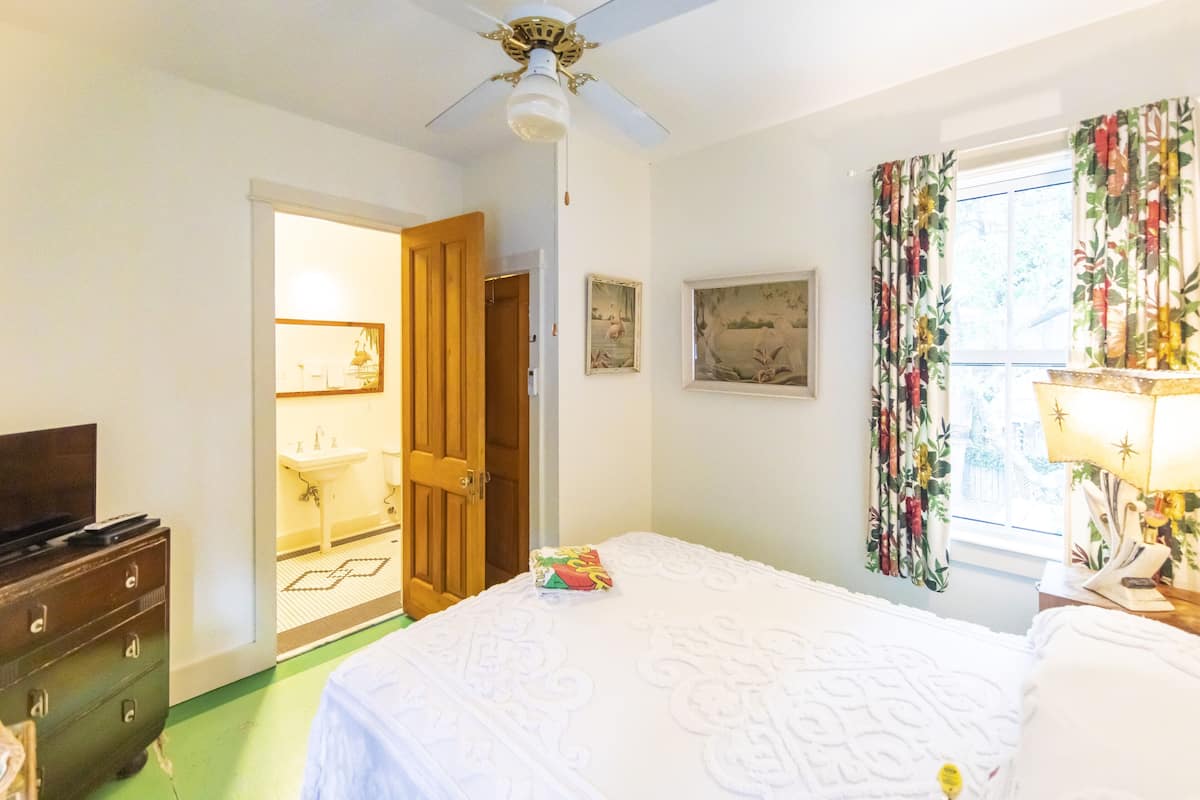 A bedroom with a bed, a dresser, a ceiling fan, a window with curtains, and a door leading to a bathroom.