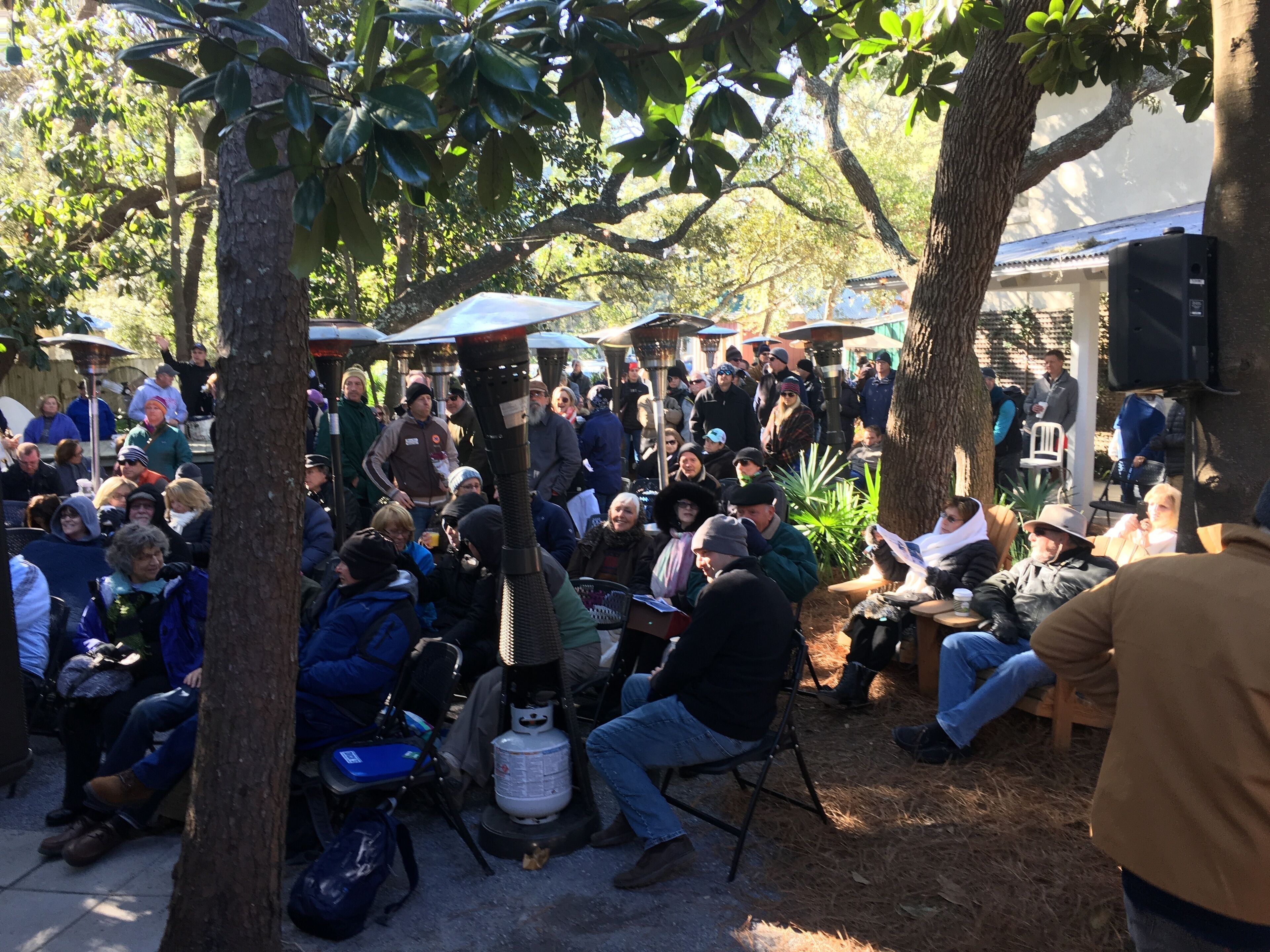 Een levendige openluchtbijeenkomst, mensen zitten onder parasols, omgeven door bomen.