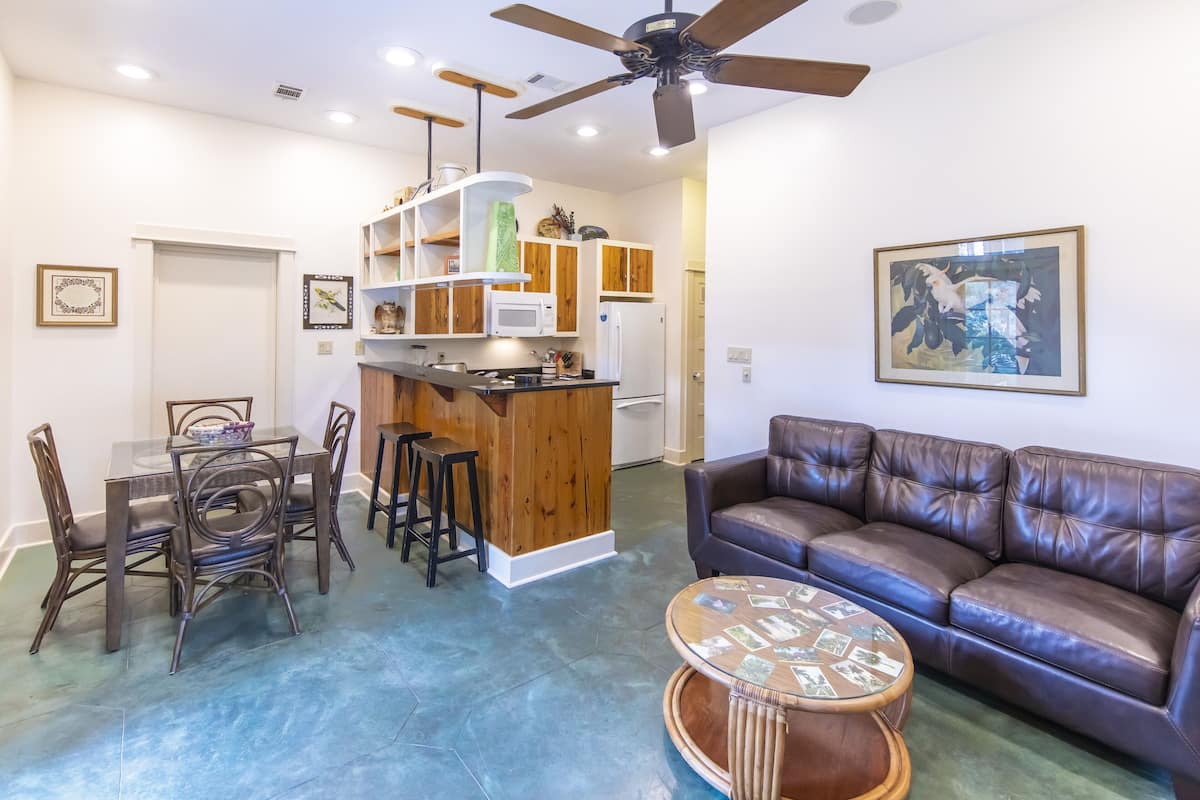 A modern living room with a brown leather sofa, a glass-top dining table with chairs, a kitchen area with wooden cabinets, and a ceiling fan.