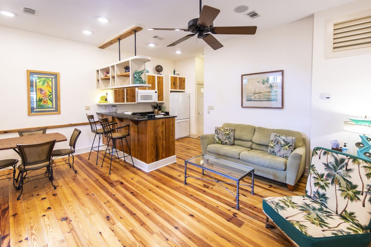 A modern living space with a wooden floor, a dining area with chairs, a sofa, a coffee table, a ceiling fan, and kitchen cabinets.