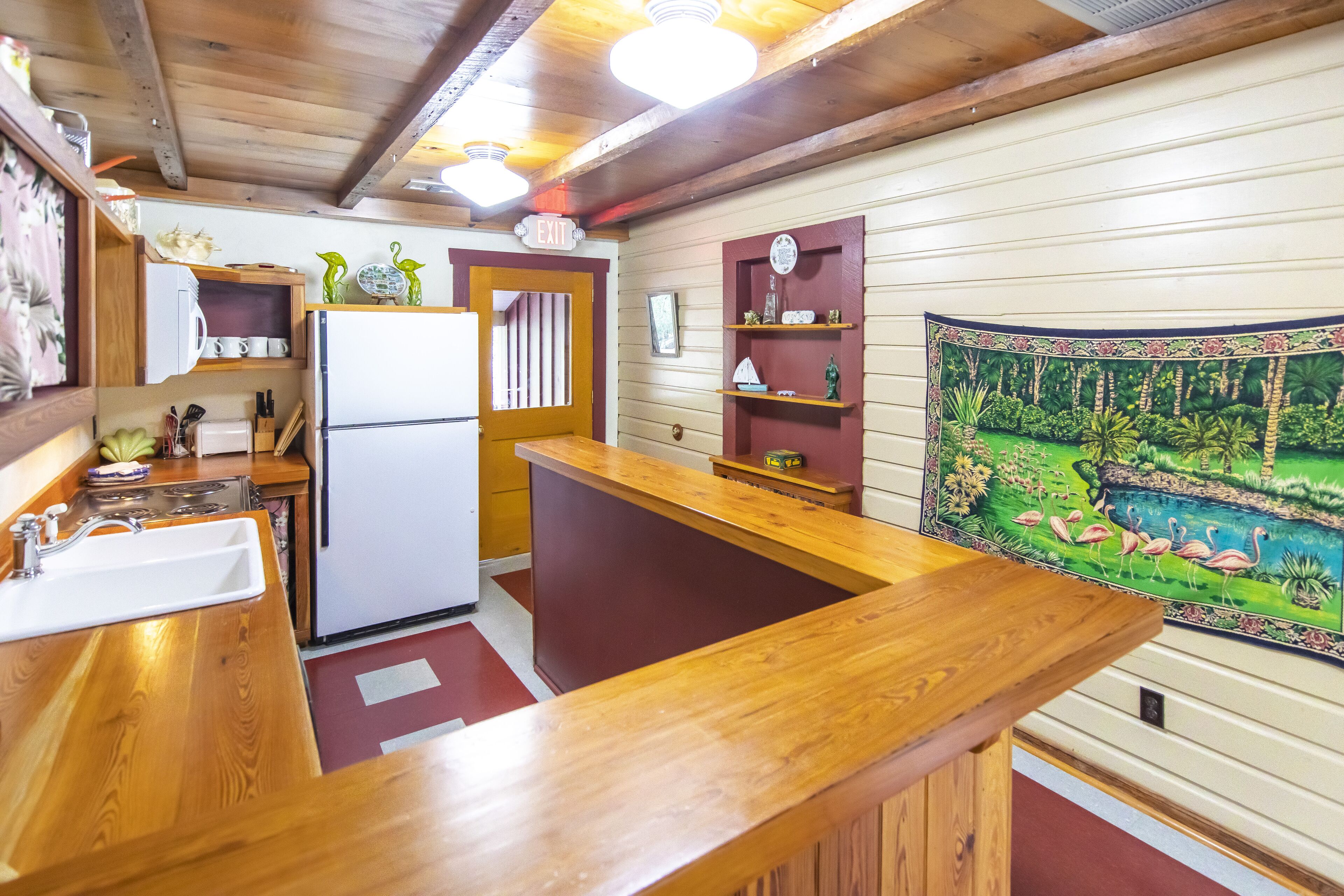 Une cuisine compacte dotée de plans de travail en bois, d’un évier, d’un réfrigérateur et d’une fresque représentant un paysage tropical.