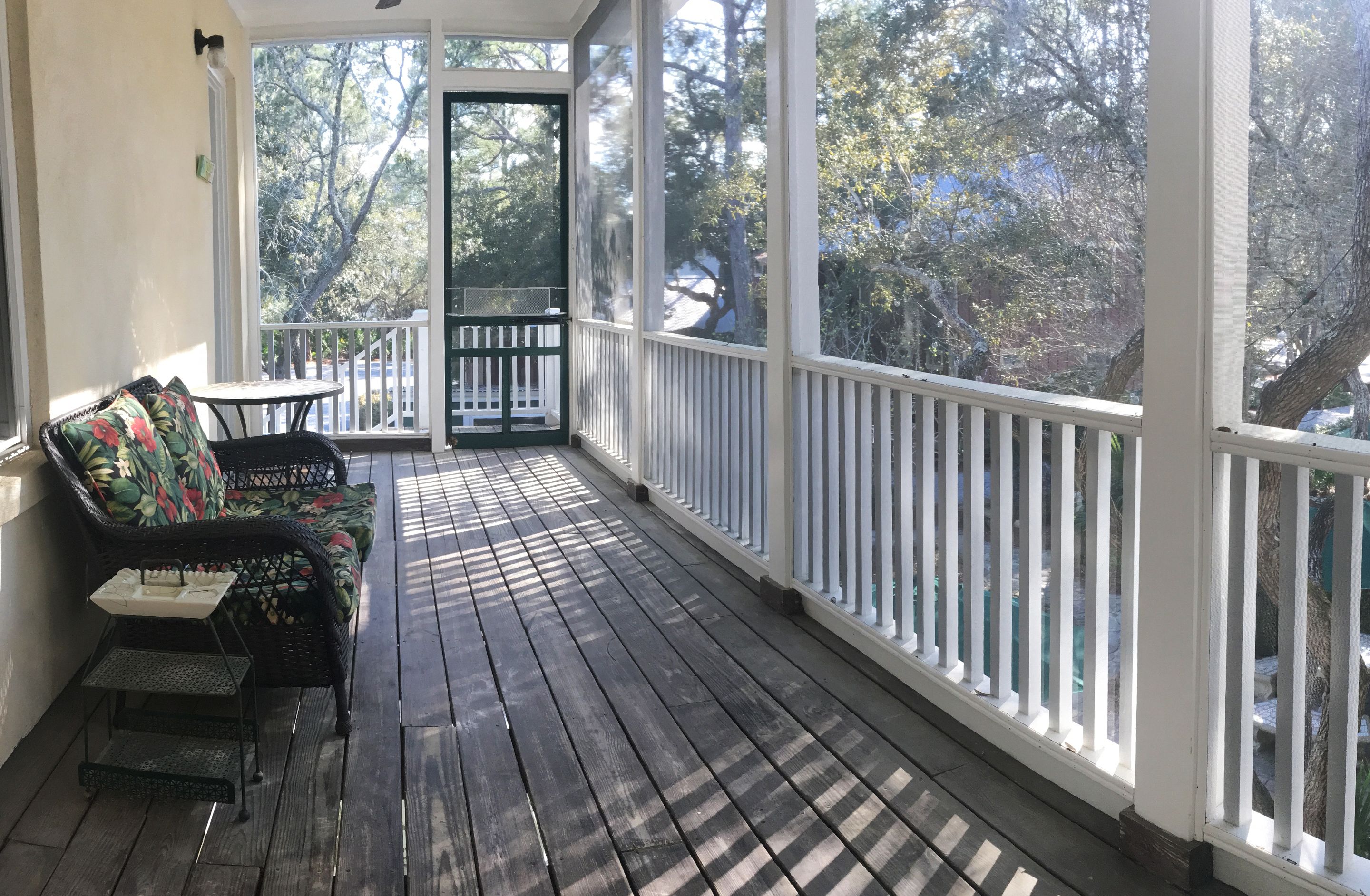 A covered porch with a wooden floor, a black wicker sofa with floral cushions, and a small table.