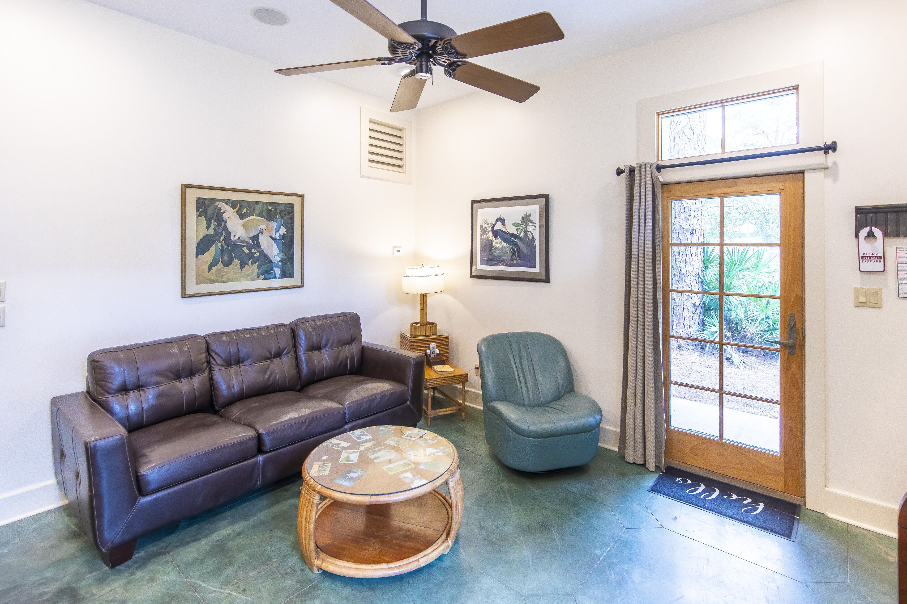 A living room with a brown leather sofa, a blue armchair, a round coffee table, a ceiling fan, and two framed pictures on the wall.