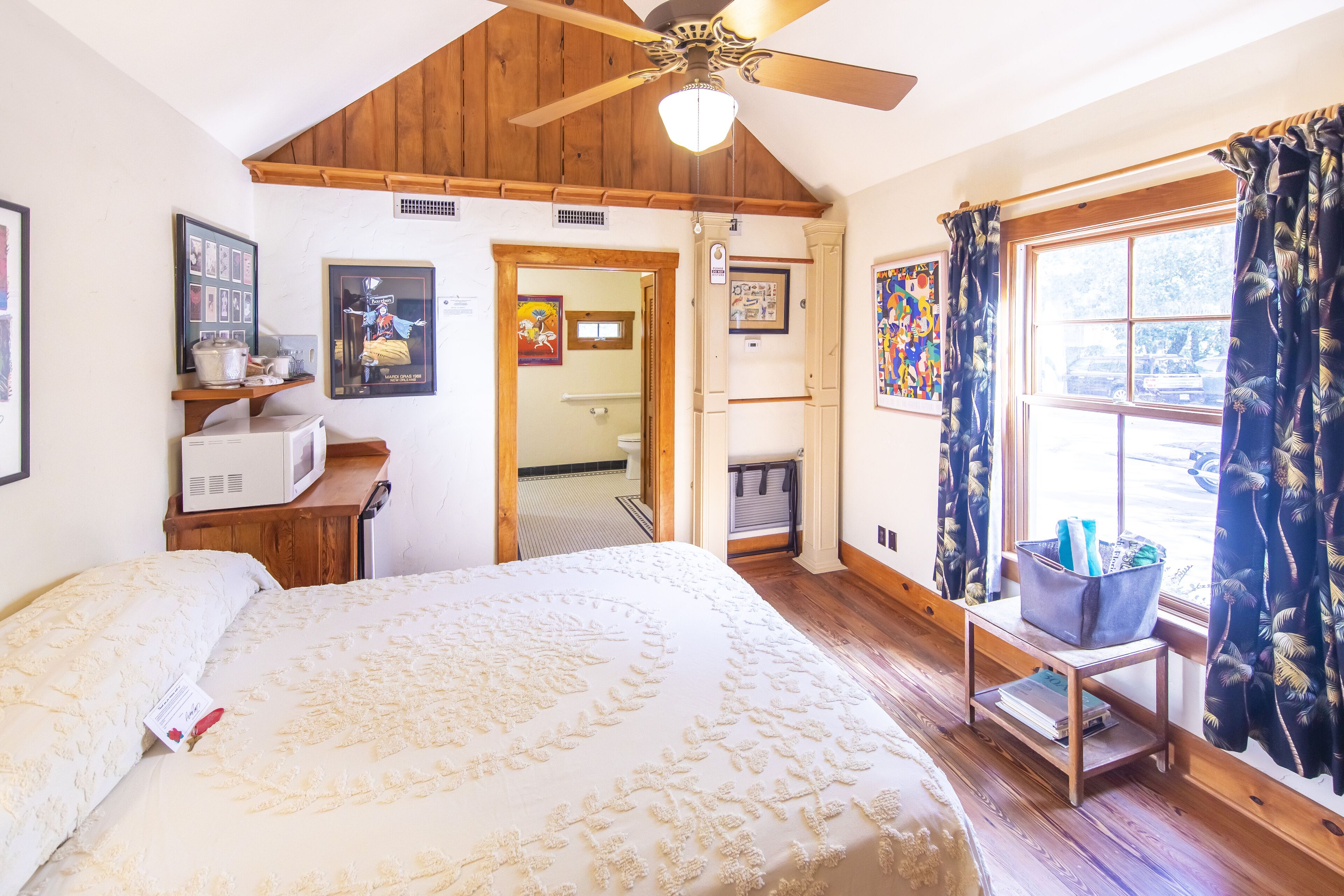 A bedroom with a large bed, wooden ceiling, a ceiling fan, a window with curtains, a small table with a basket, and a microwave on a wooden shelf.