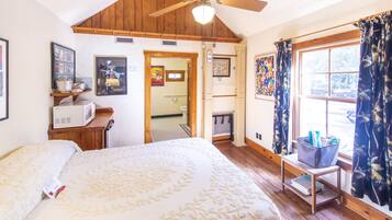 A bedroom with a large bed, wooden ceiling, a ceiling fan, a window with curtains, a small table with a basket, and a microwave on a wooden shelf.