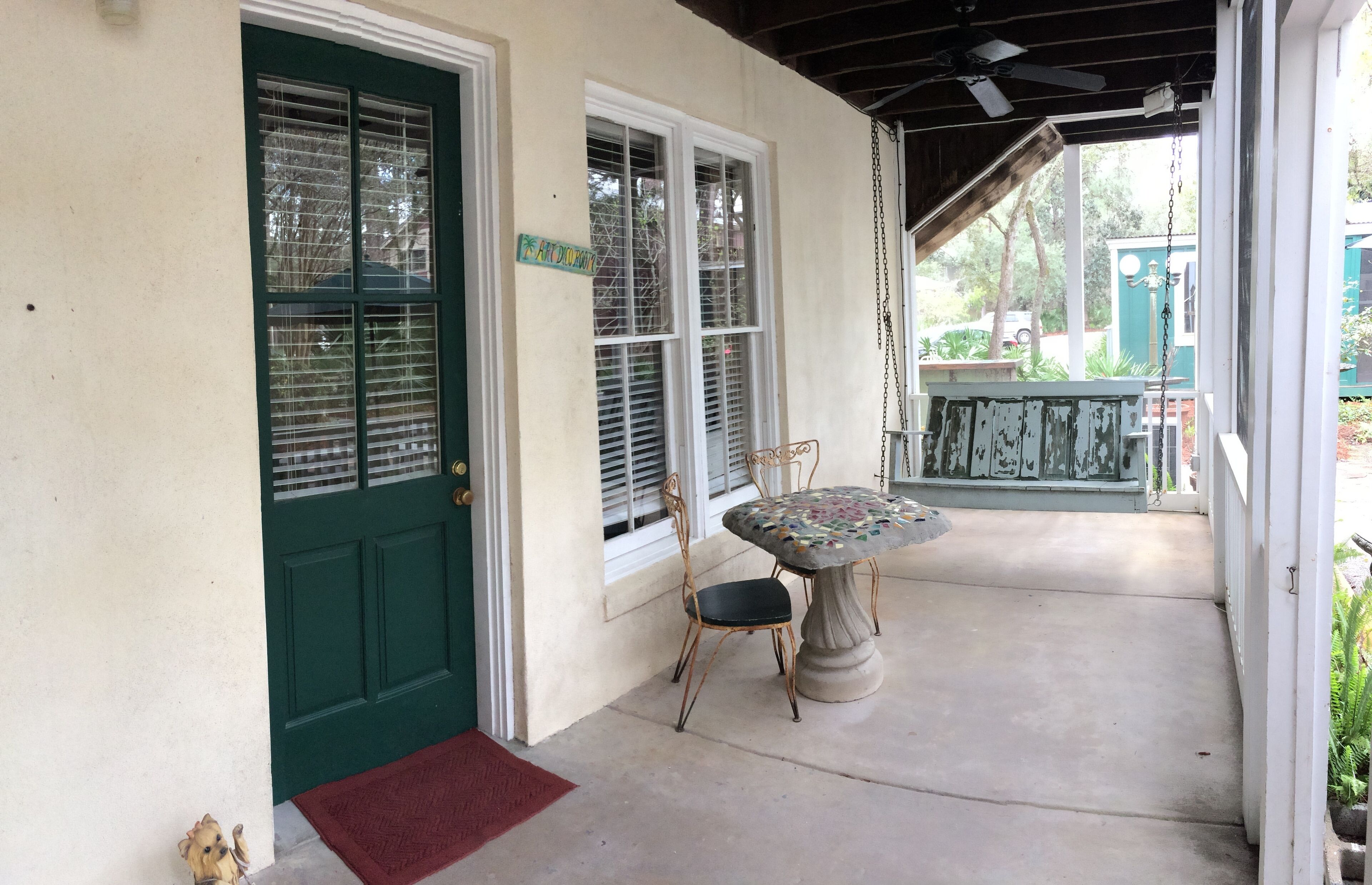 A covered porch with a green door, a small table with chairs, and a red mat at the entrance.