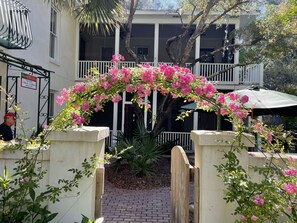 A courtyard with a wooden archway, pink flowering plants, and a white building with balconies.