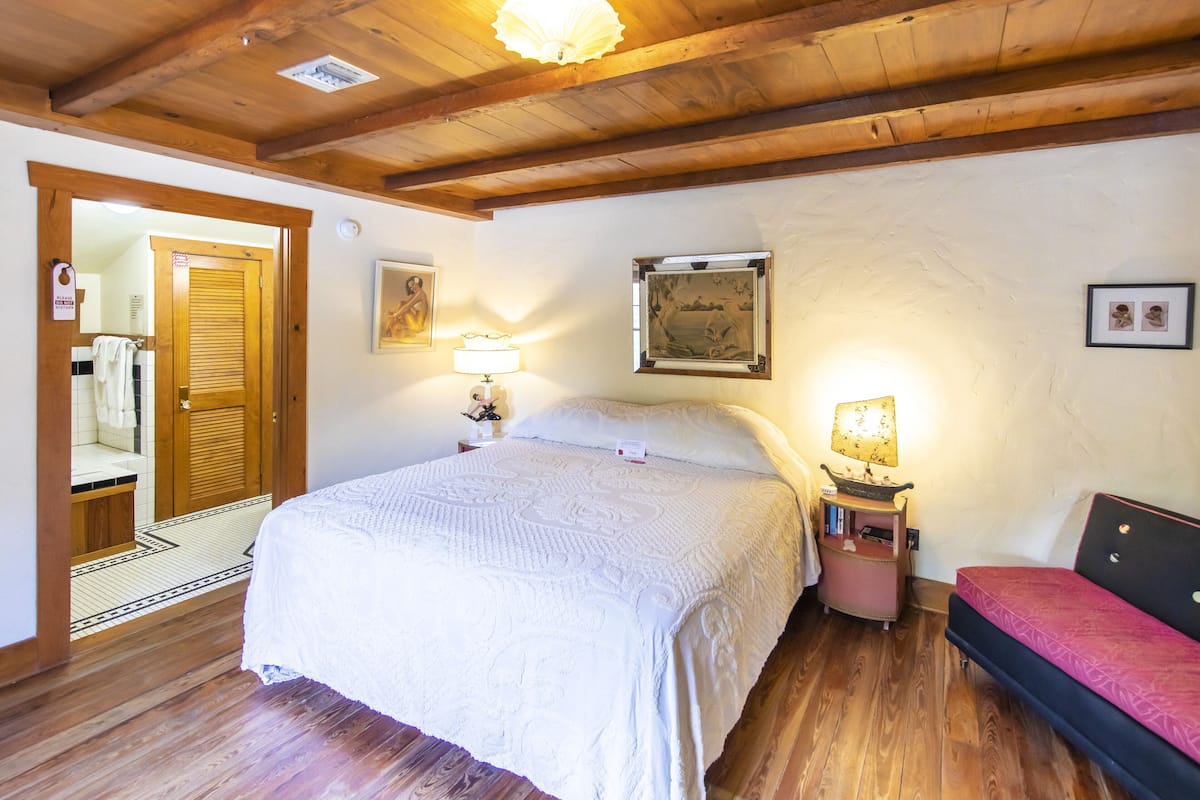 A bedroom with a large bed, wooden ceiling beams, a nightstand with a lamp, a small table with books, and a red bench.