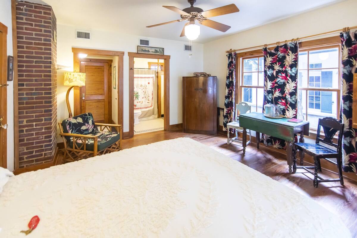 A bedroom with a large bed, a wooden desk, a chair, a lamp, and a window with curtains.