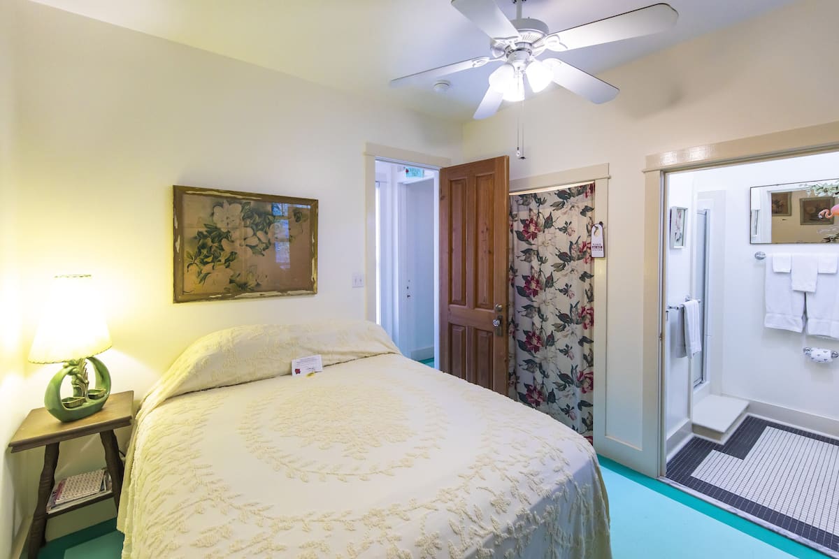 A bedroom with a bed, a nightstand, a ceiling fan, a door leading to a bathroom, and a floral curtain.
