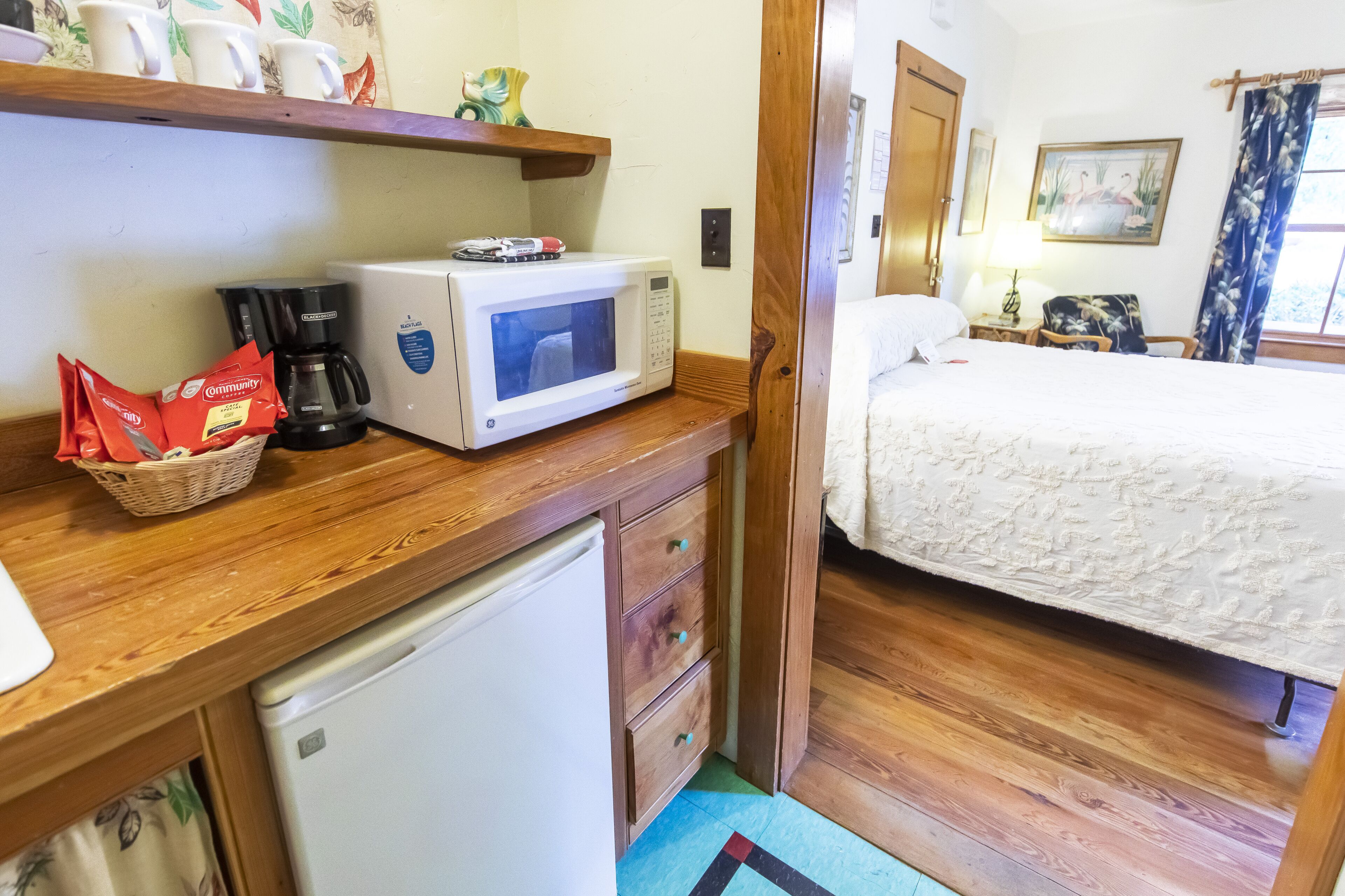 Una kitchenette compacta con microondas, cafetera y refrigerador. Una pequeña cesta con paquetes rojos está sobre la encimera.