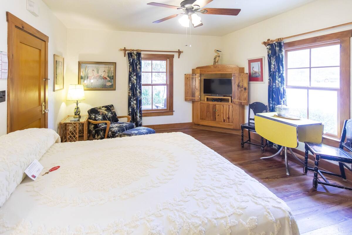 A bedroom with a large bed, a wooden desk with a chair, a television, and a ceiling fan.