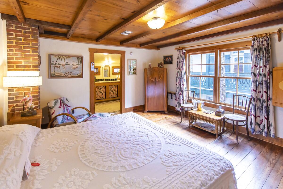 A cozy room with a large bed, wooden ceiling, a dining area with a table and chairs, and a view through a window with curtains.
