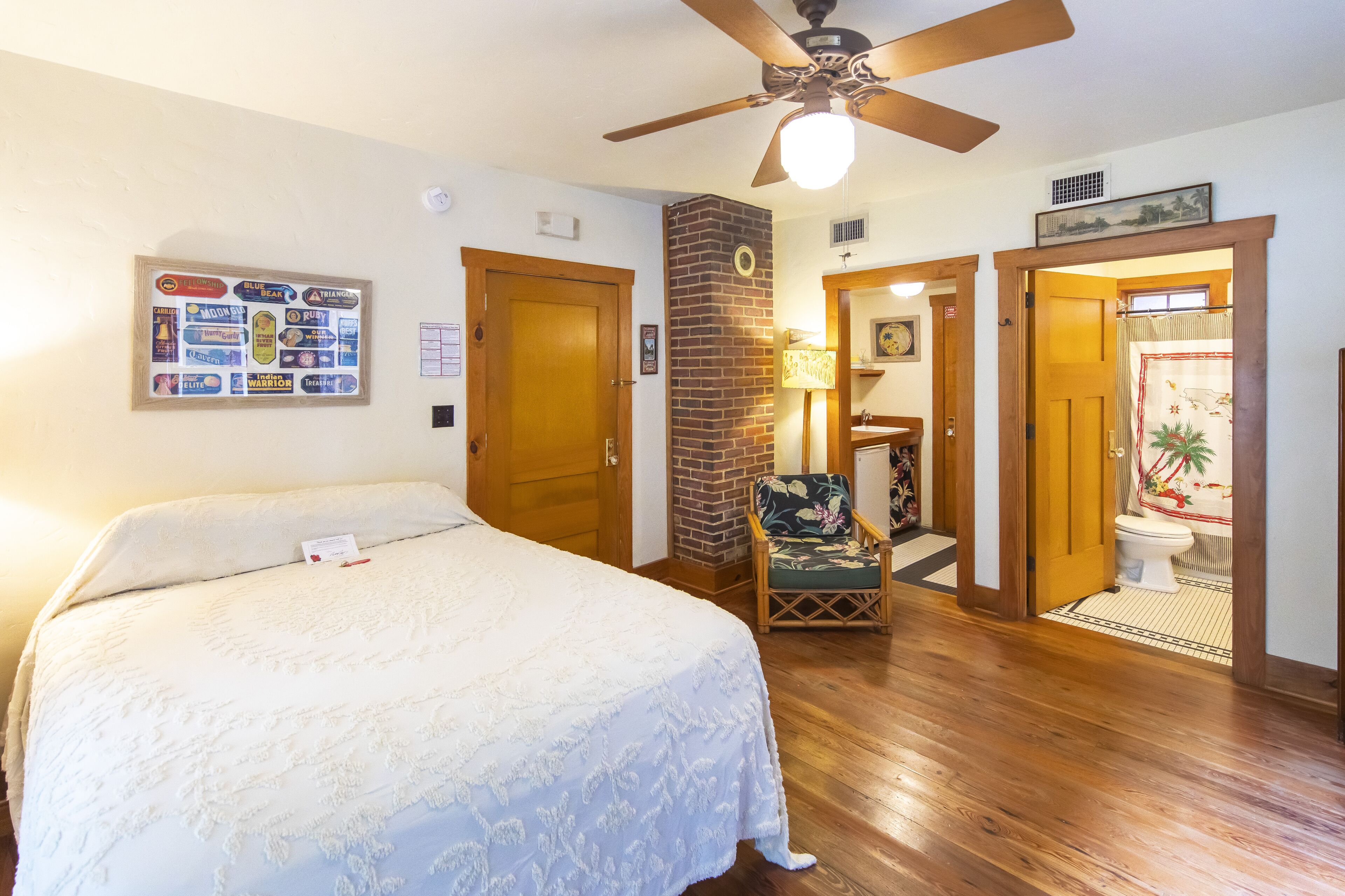 A bedroom with a large bed, a ceiling fan, a wooden floor, a brick wall, and a bathroom visible through an open door.