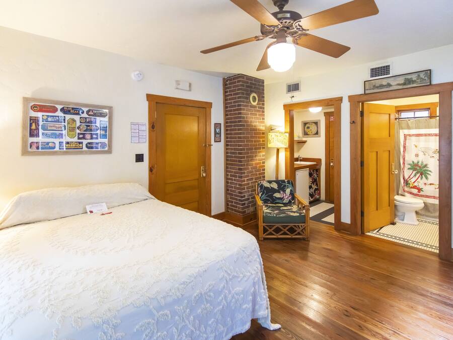 A bedroom with a large bed, a ceiling fan, a wooden floor, a brick wall, and a bathroom visible through an open door.