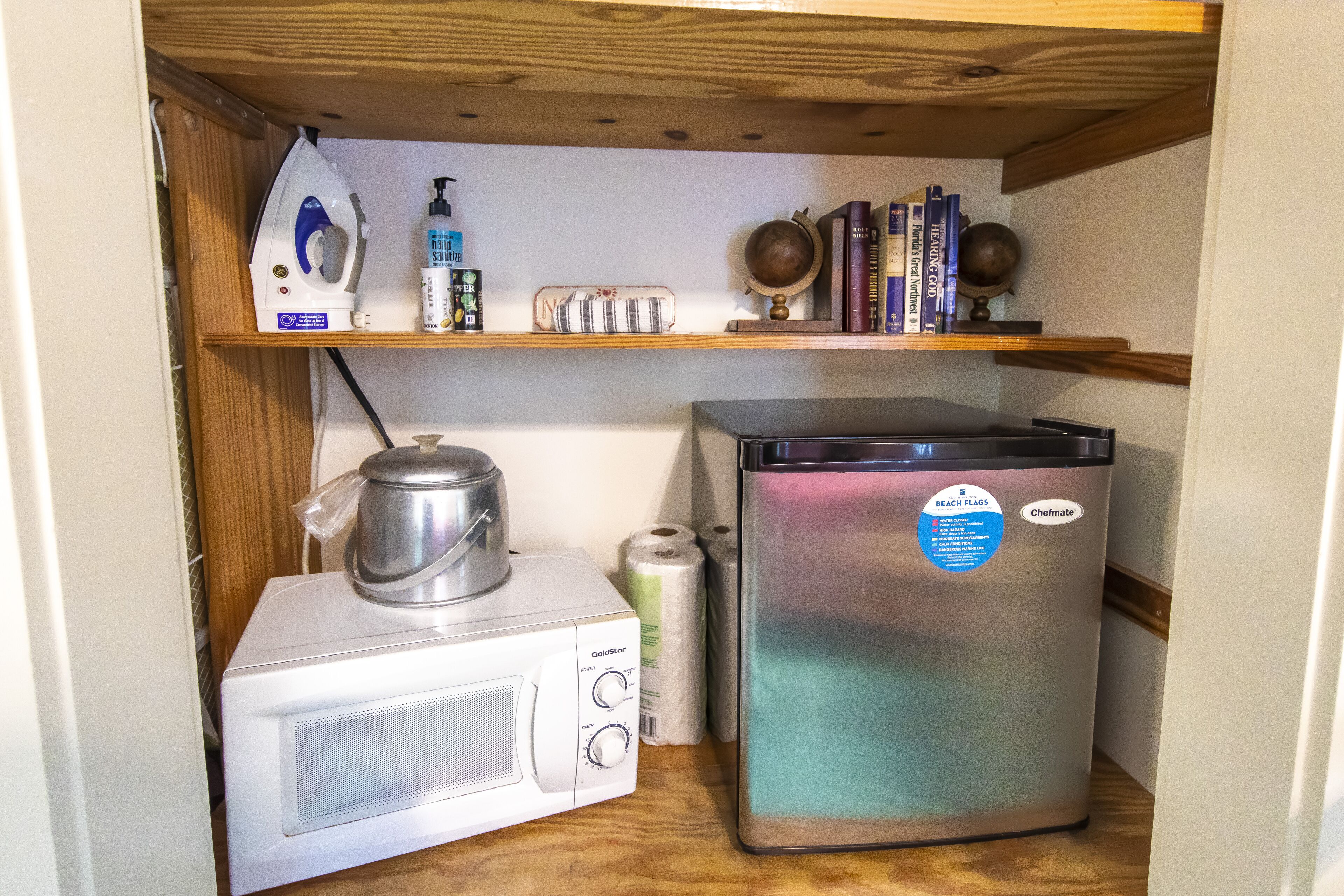 Una kitchenette compacta con microondas, hervidor de agua, refrigerador y una estantería con libros y utensilios de cocina.