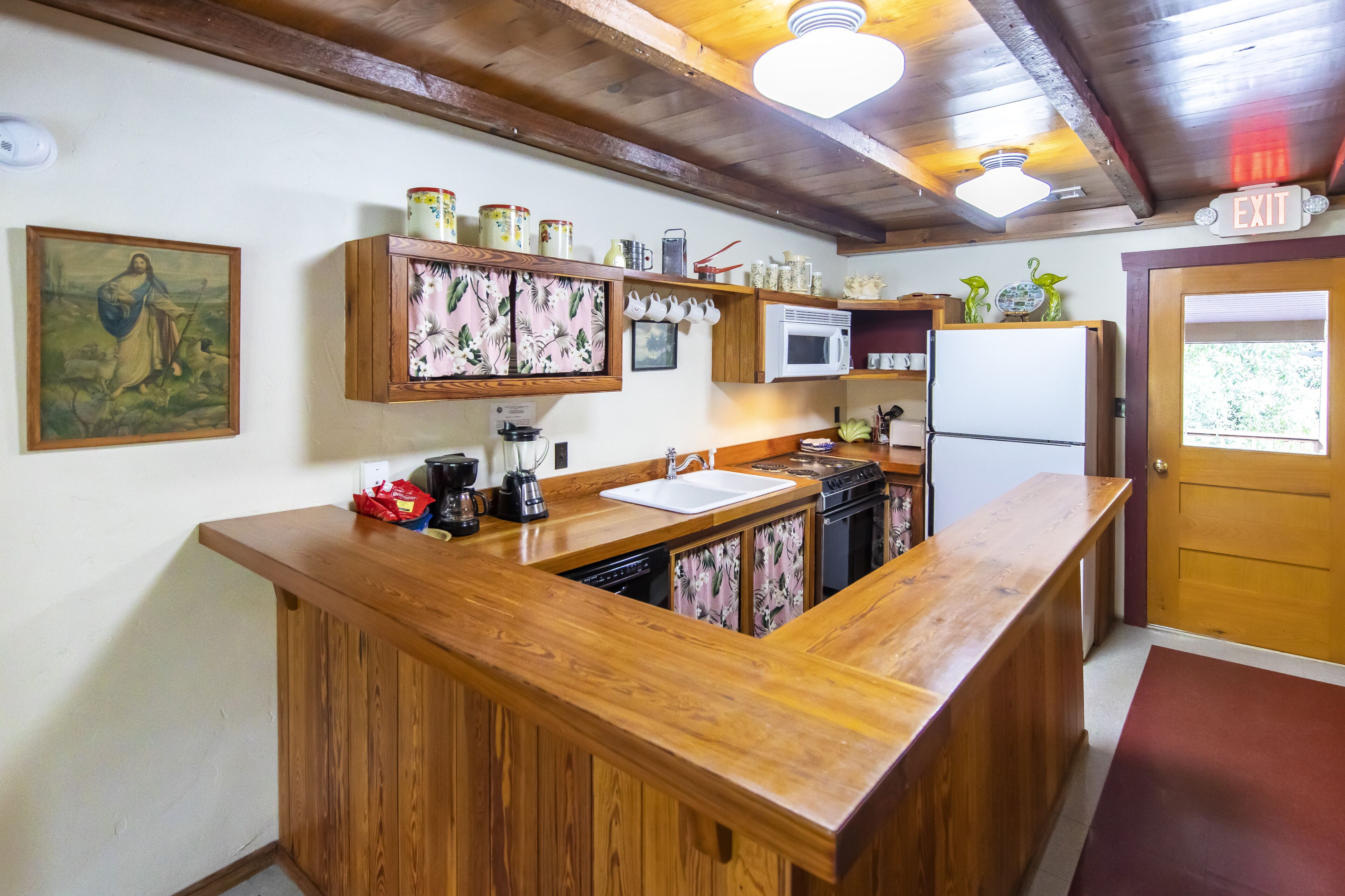 A kitchen with wooden countertops, a microwave, a refrigerator, and a sink. There is a painting on the wall and a shelf with jars and kitchen items.