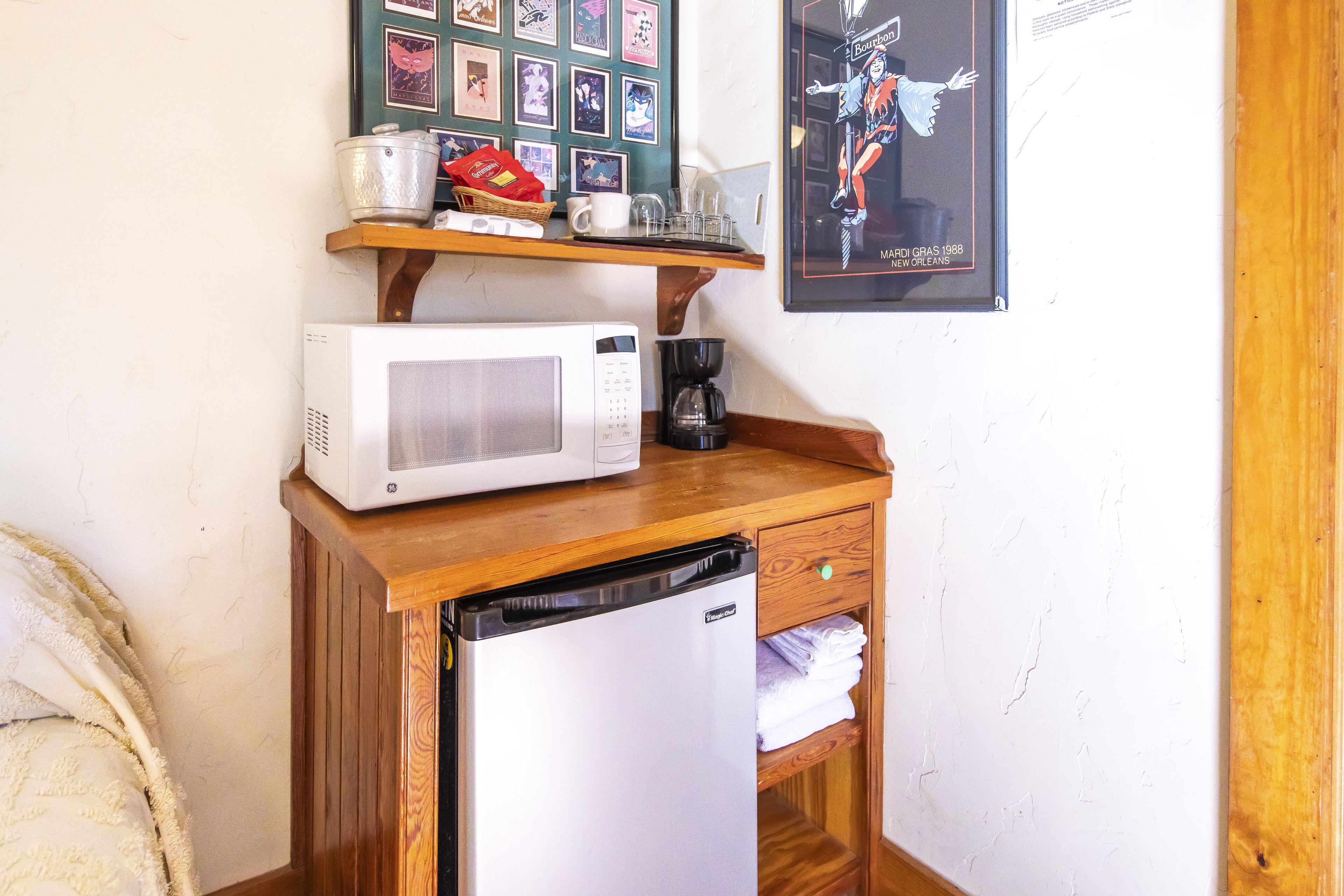 A compact kitchenette with a microwave, coffee maker, and a small refrigerator. A wooden shelf above holds various kitchen items.