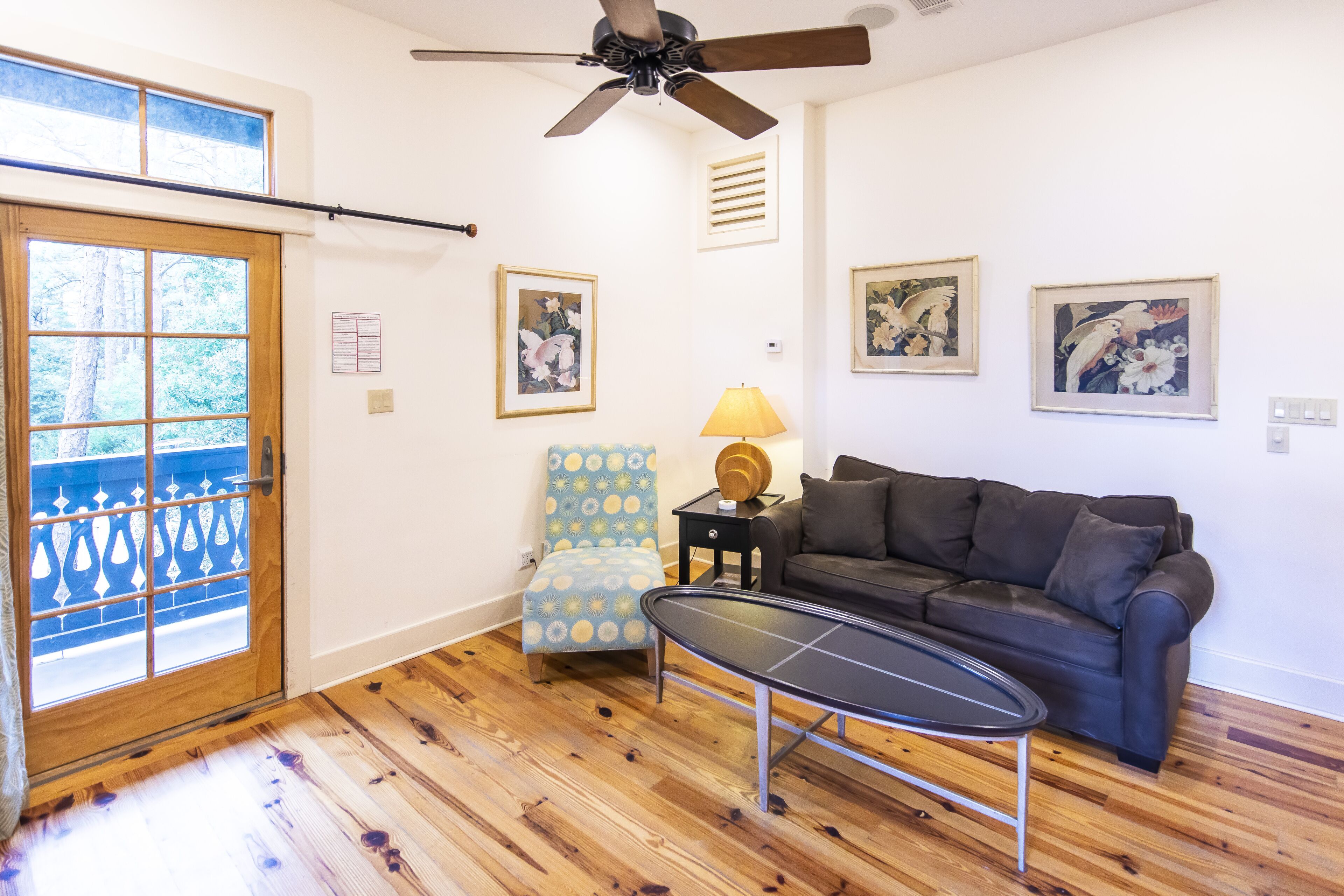 A room with a wooden floor, a black leather sofa, a blue patterned chair, a small table, and a ceiling fan.
