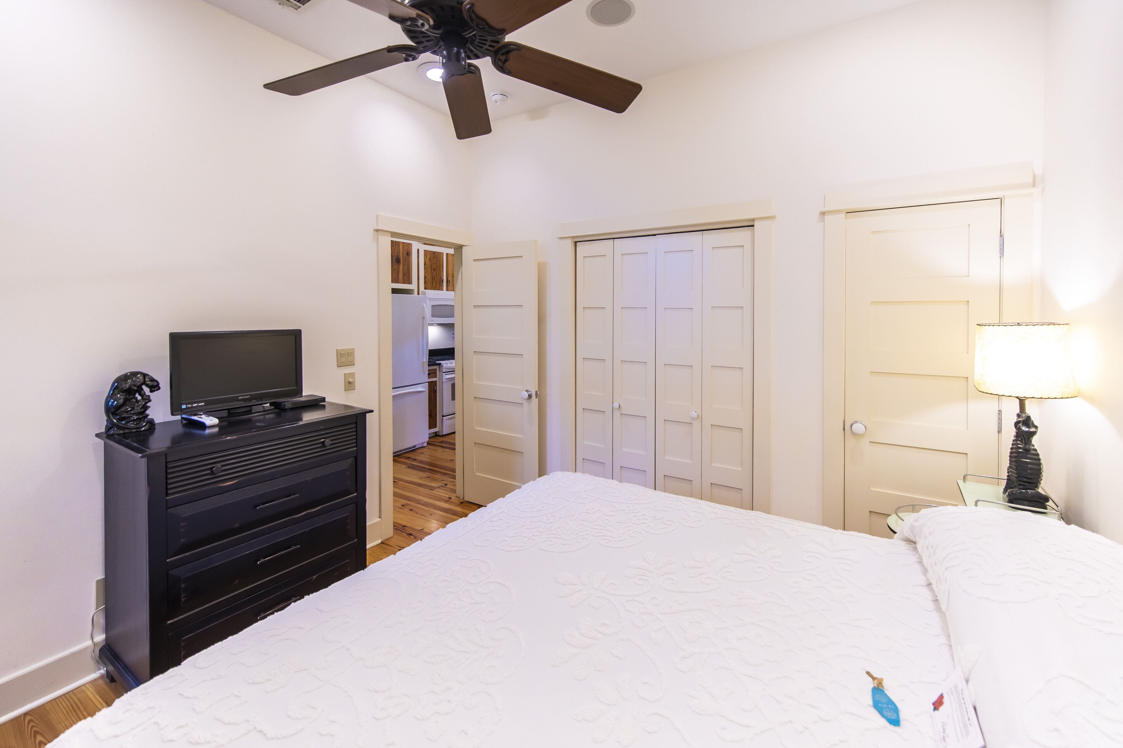 A bedroom with a bed, a TV on a dresser, a ceiling fan, and a door leading to a kitchen.