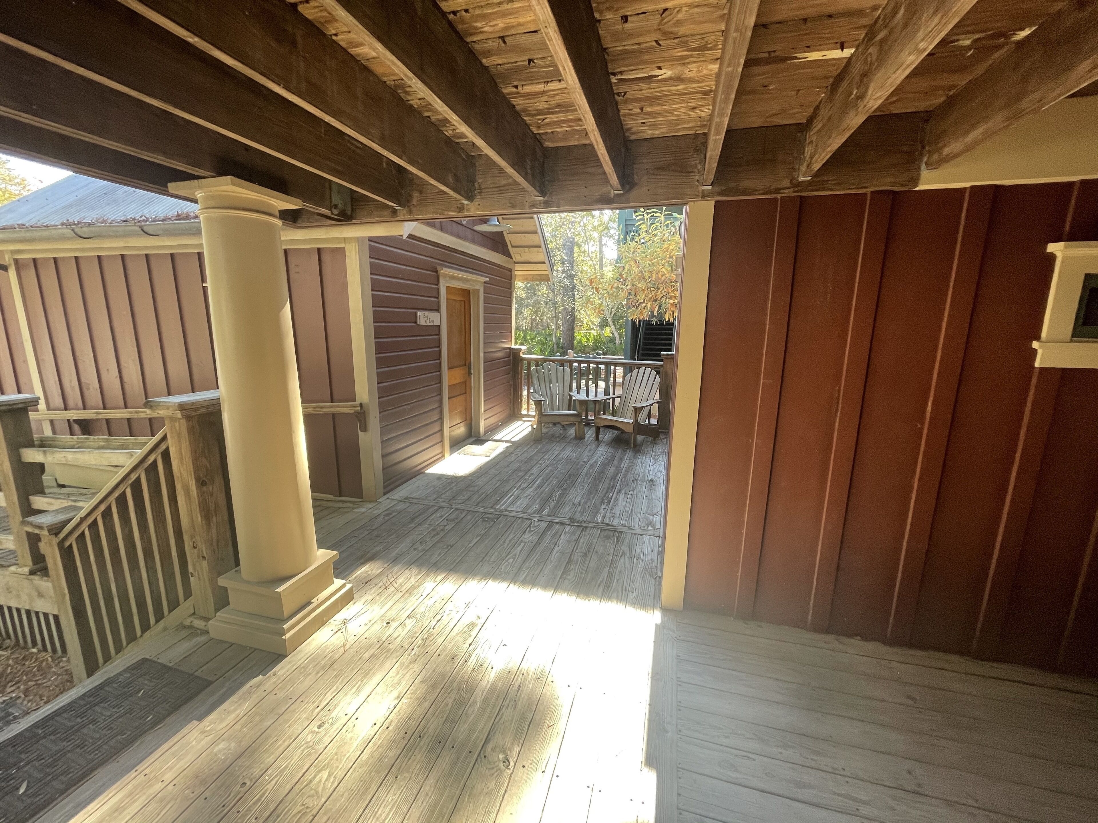 A covered wooden deck with a staircase leading to an upper level, a small outdoor seating area, and a view of the surrounding trees.