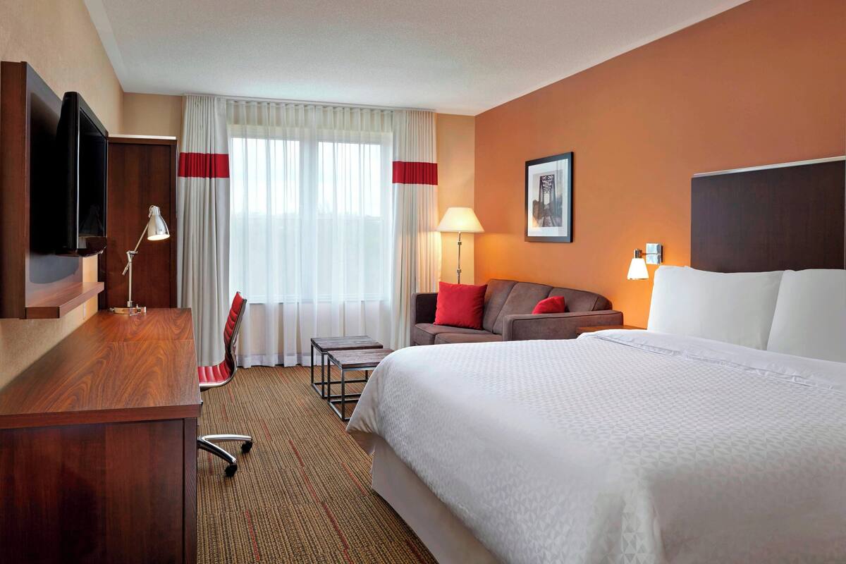 A hotel room with a large bed, a desk with a flat-screen TV, a sofa with red cushions, a lamp, and a framed picture on the wall.