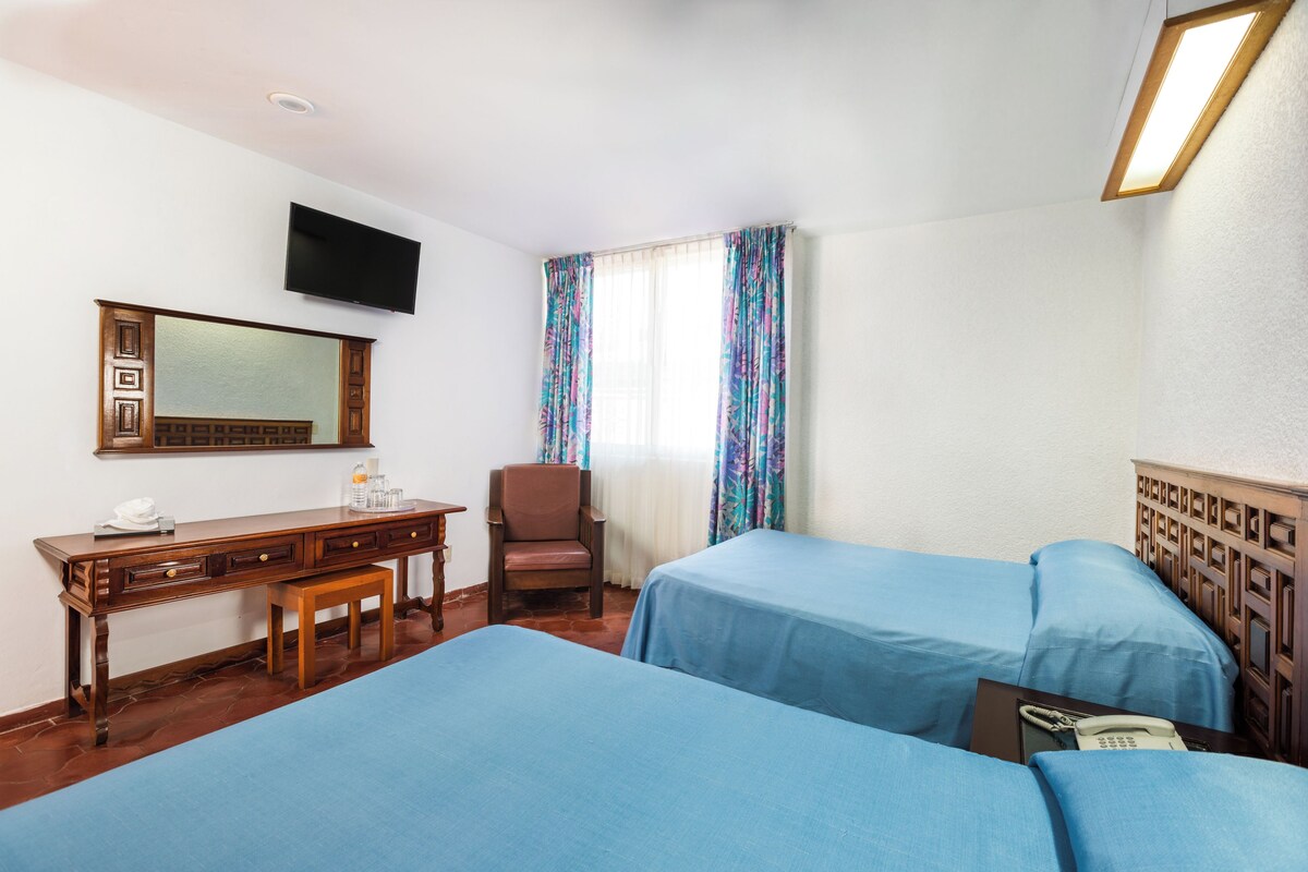 A hotel room with two beds, a desk, a chair, a TV, and a window with curtains.