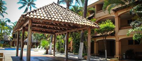 A covered pavilion with a tiled roof, palm trees, and a pool in the background.