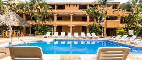 A swimming pool with lounge chairs, palm trees, and a building with multiple balconies.