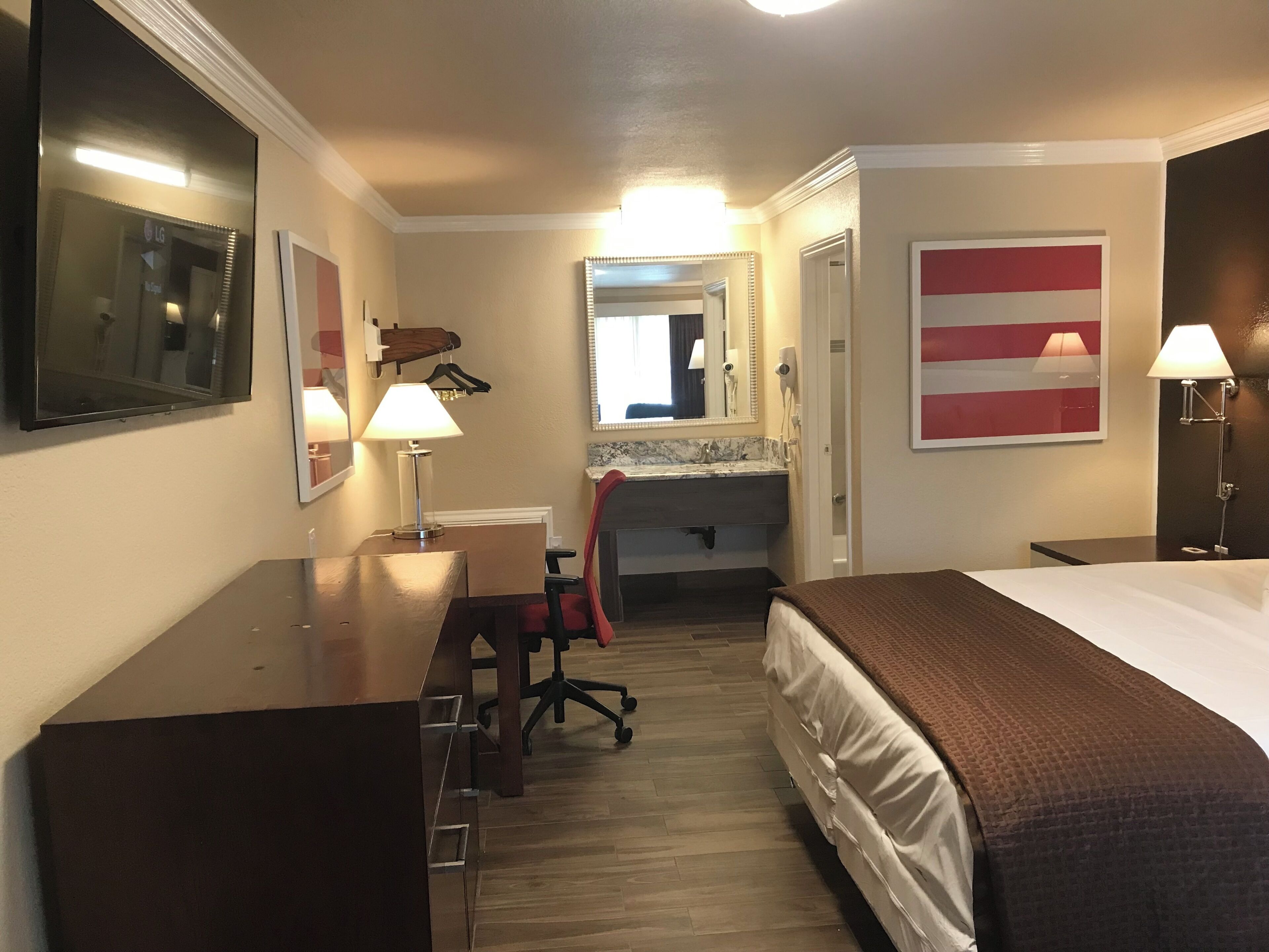 A hotel room with a bed, desk, chair, mirror, and a bathroom with a sink and mirror.