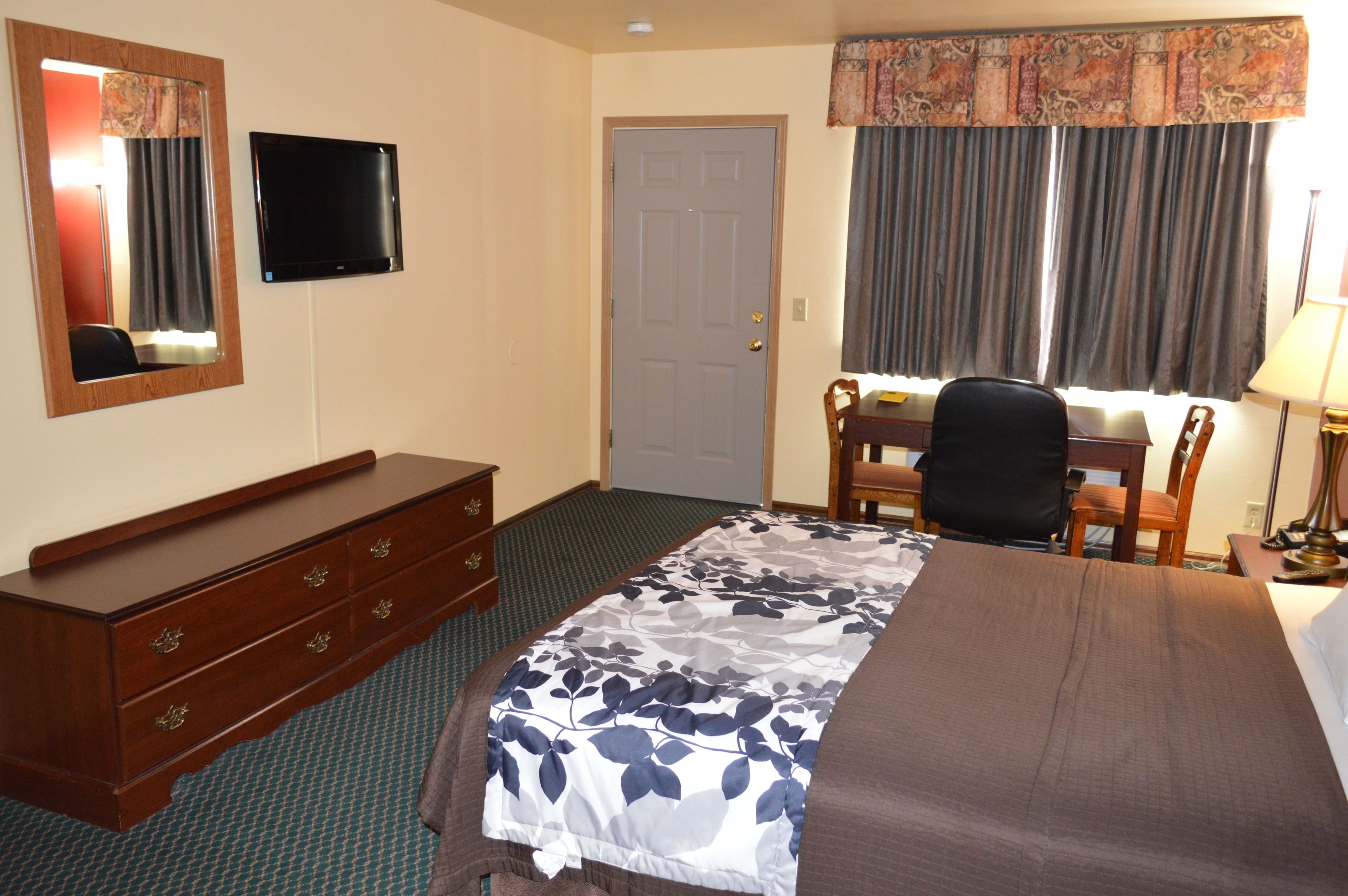 A hotel room with a bed, a dresser, a desk with a chair, a television, and a window with curtains.