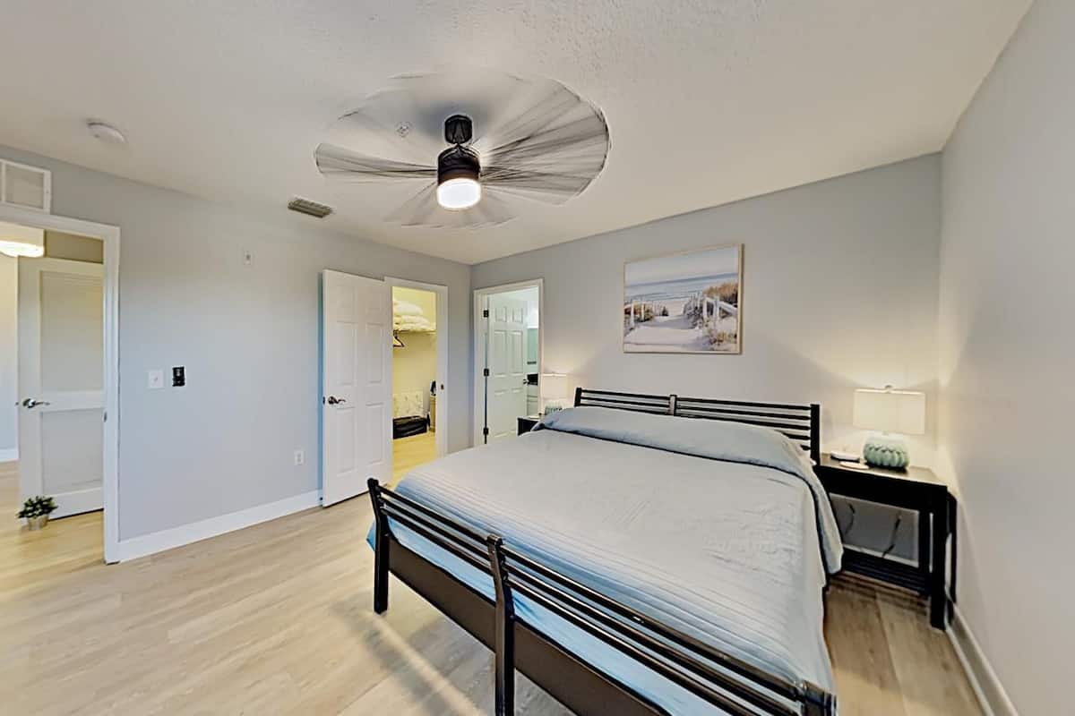 Une chambre à coucher comprenant un lit, un ventilateur de plafond, une table de chevet avec une lampe et une porte donnant sur une autre pièce.