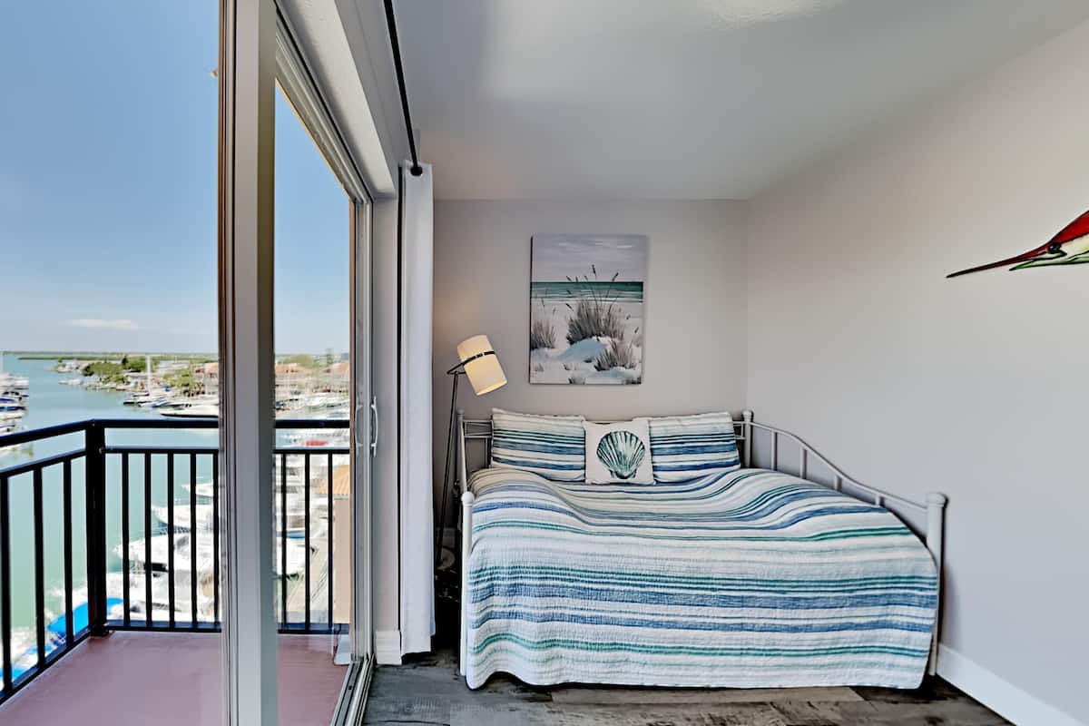 Un balcon donnant sur des bateaux et la verdure, un lit avec une literie à rayures et des oreillers, une lampe sur pied et un tableau accroché au mur.