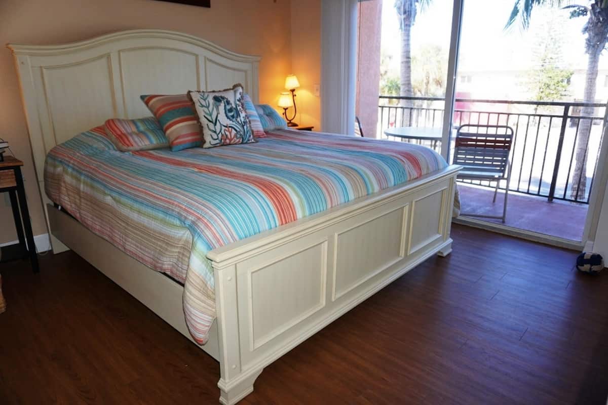Un lit avec une couvre-lit à rayures et plusieurs oreillers, une table de chevet avec une lampe, et une vue sur un balcon avec une chaise.