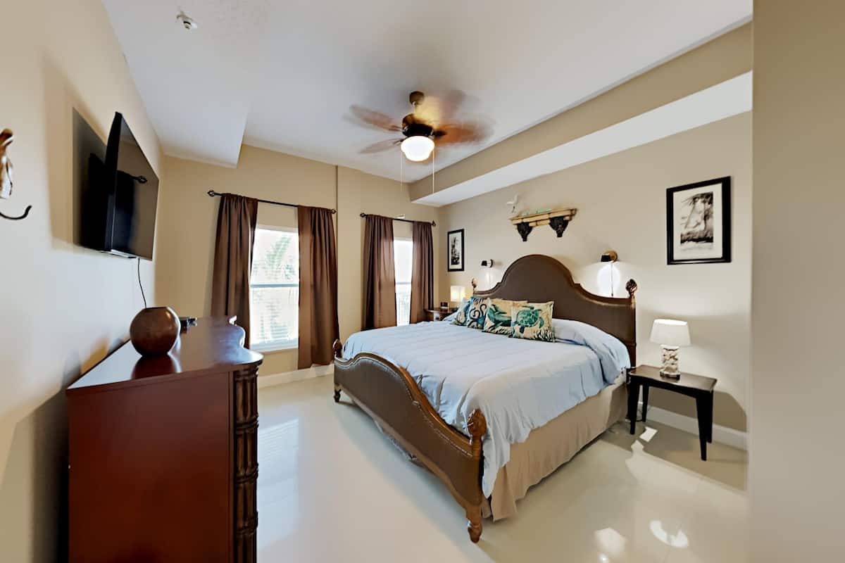 Une chambre à coucher comprenant un lit, un ventilateur de plafond, une télévision, une commode et une fenêtre avec des rideaux.