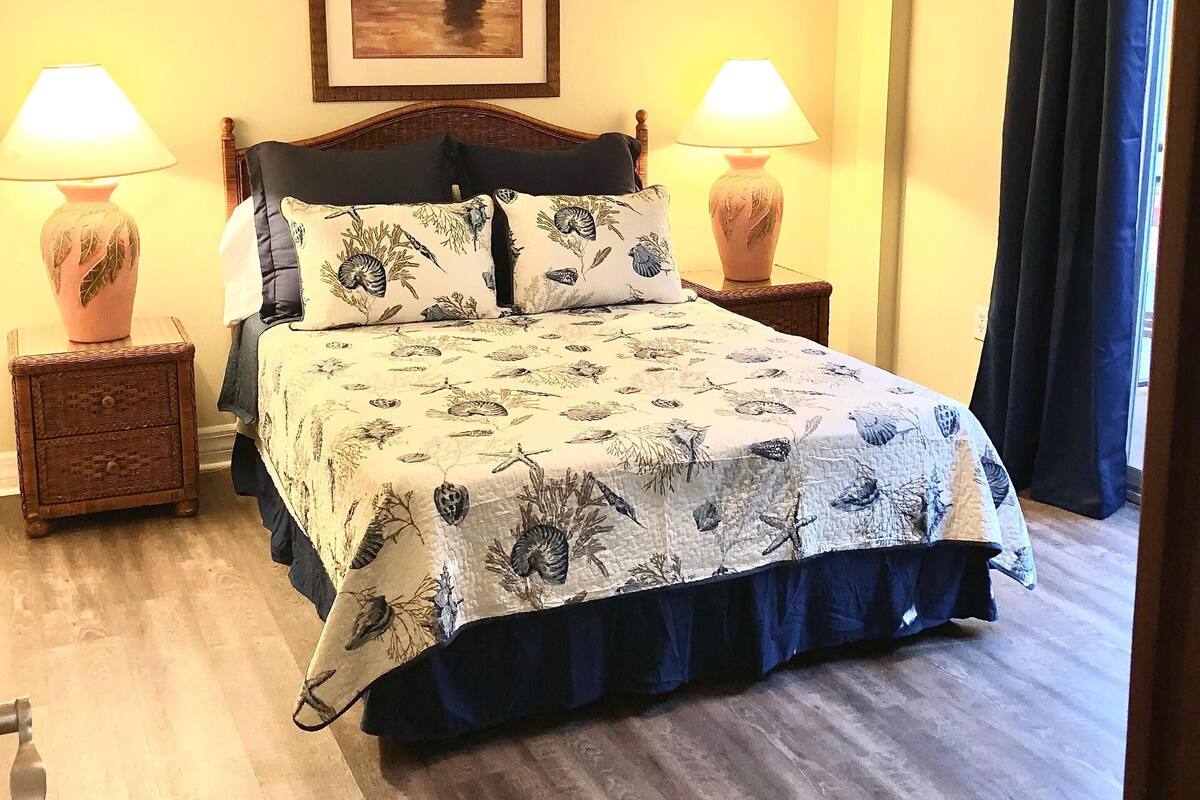 Un lit bien fait, recouvert d’une couvre-lit à motifs floraux, deux lampes de chevet, un ventilateur de plafond et un tableau encadré au mur.