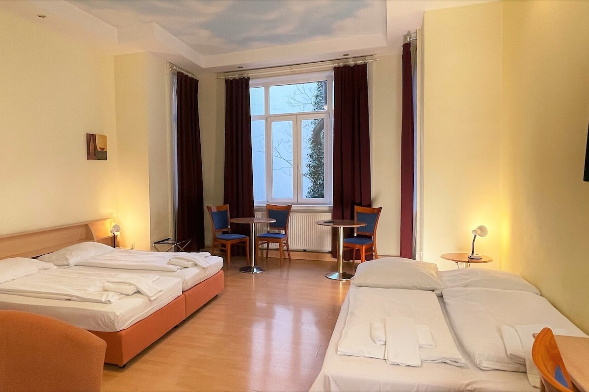 Quarto de hotel com duas camas, uma mesinha, cadeiras e uma janela com cortinas.