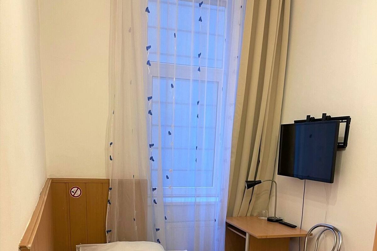 Uma cama de solteiro com roupa de cama branca, cabeceira de madeira, uma escrivaninha com cadeira, uma televisão e uma janela com cortinas.