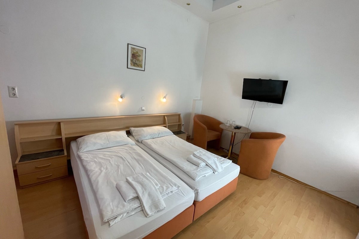 Quarto de hotel com uma cama de casal, uma mesinha, duas cadeiras, uma TV fixada na parede e um quadro na parede.