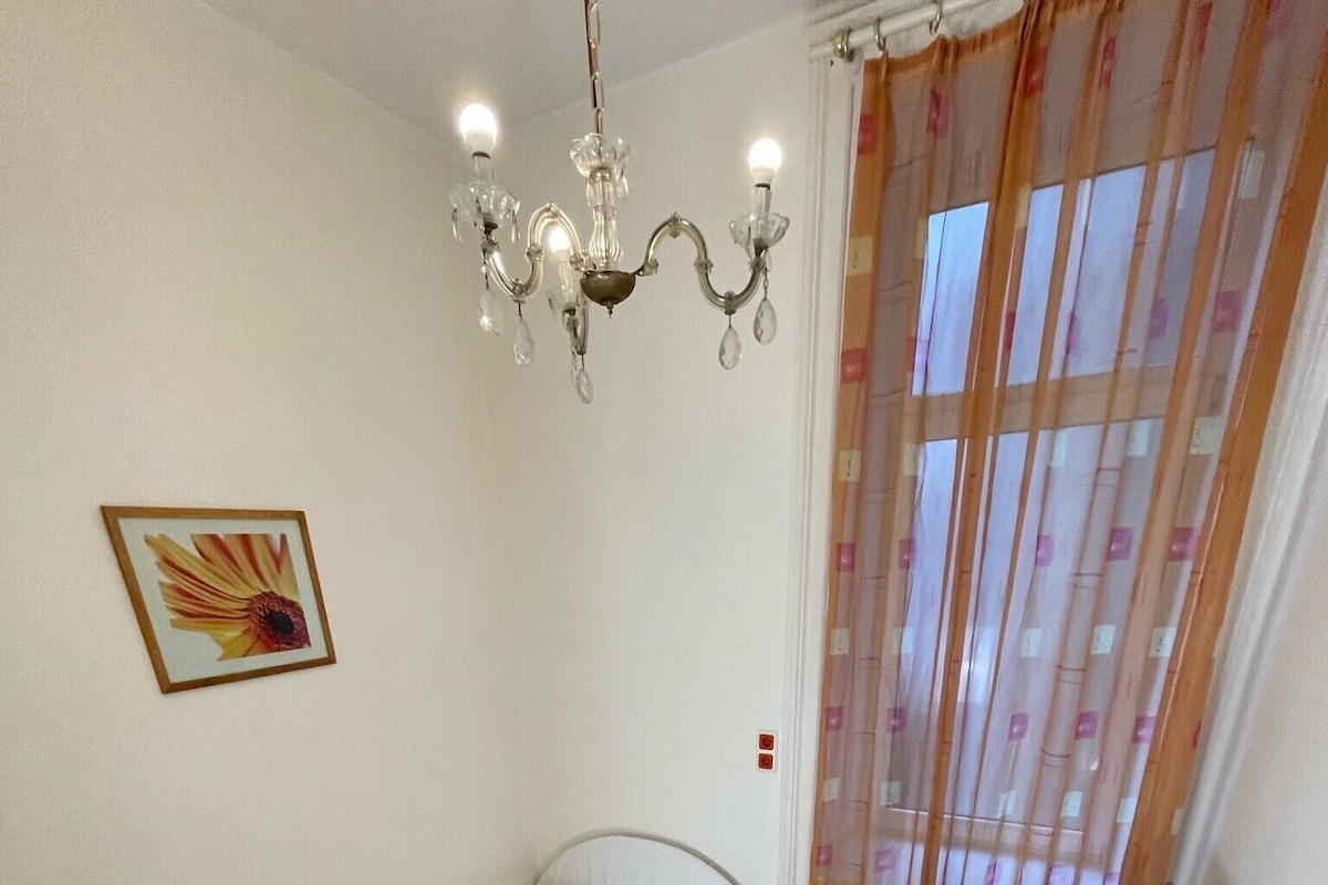 Quarto pequeno com uma cama de solteiro, escrivaninha, cadeira, lustre e janela com cortinas.