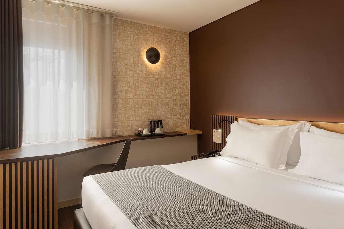 A hotel room with a bed, a desk, a chair, a coffee maker, and a wall-mounted light fixture.