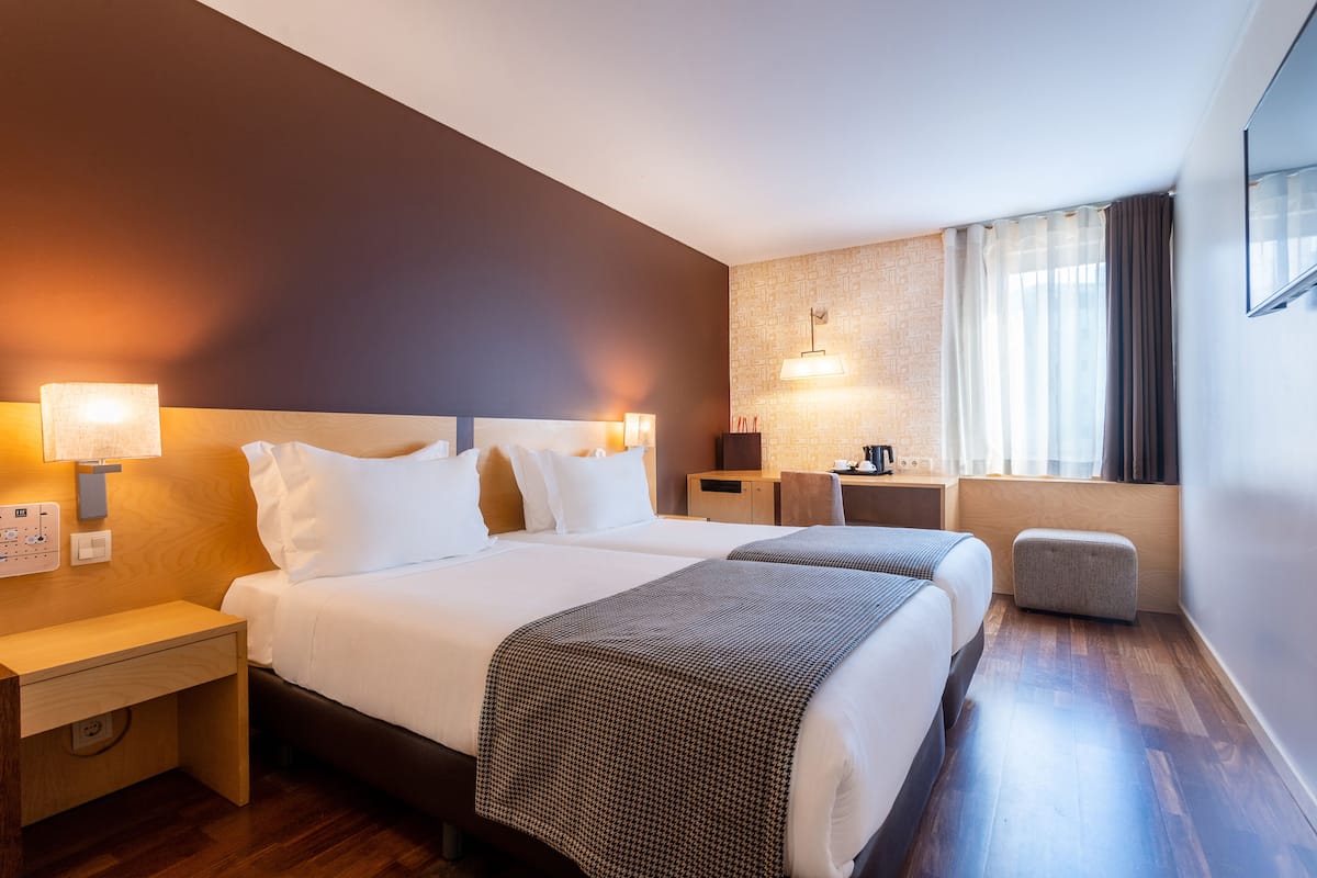 A hotel room with two beds, a desk, a chair, a lamp, and a window with curtains.