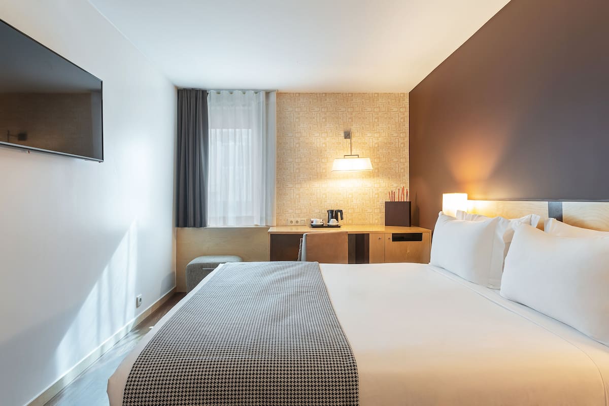 A hotel room with a large bed, a desk with a lamp, and a TV mounted on the wall.