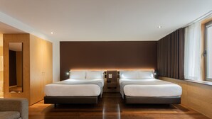Quarto de hotel moderno com duas camas, um sofá, uma janela grande com cortinas e piso de madeira.