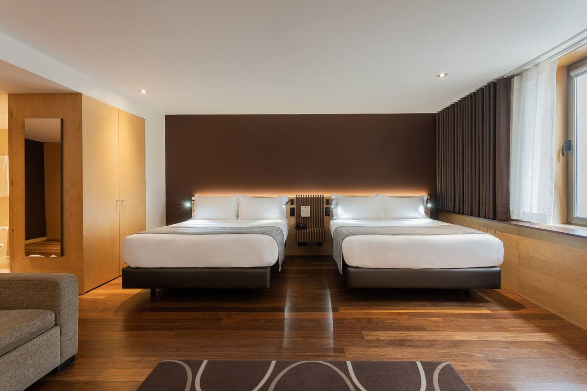 A modern hotel room with two beds, a sofa, a large window with curtains, and a wooden floor.